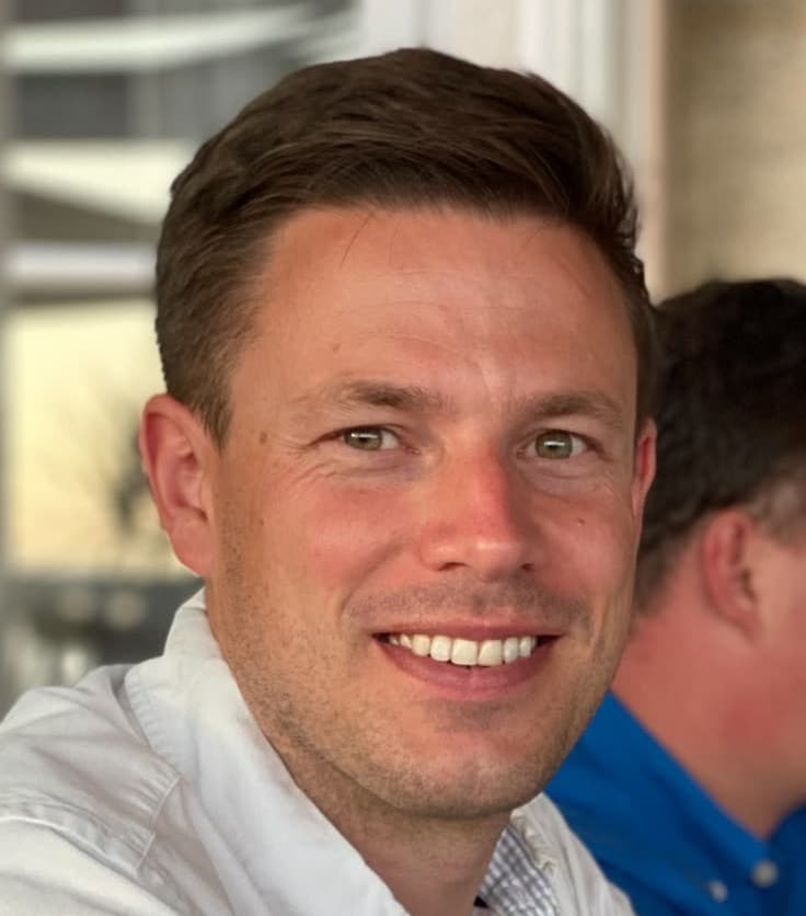 Close-up portrait of a smiling man with short brown hair wearing a white shirt.
