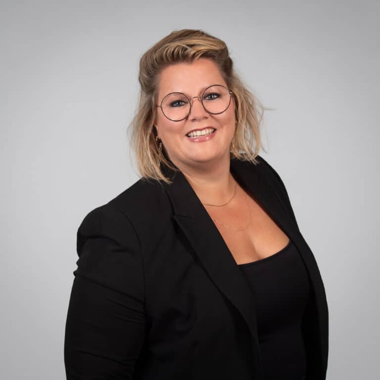 Smiling woman with round glasses and blonde hair wearing a black blazer against a gray studio backdrop.