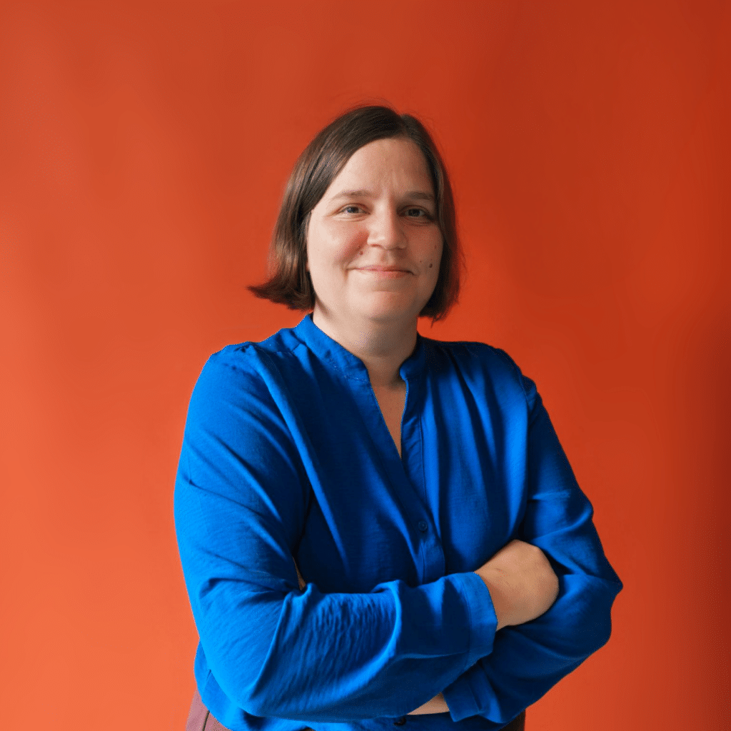 Portrait of a woman with short brown hair wearing a blue blouse, arms crossed, against an orange background.