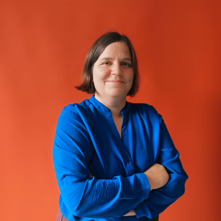Portrait of a woman with short brown hair wearing a blue blouse, arms crossed, against an orange background.