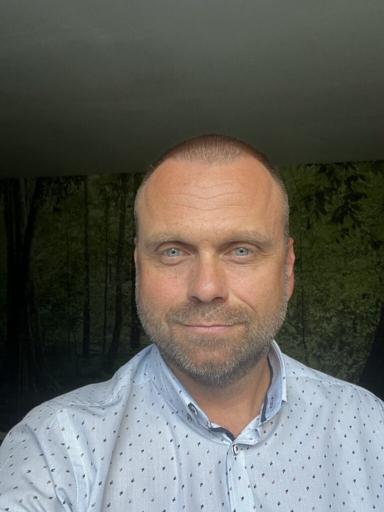 Close-up selfie of a middle-aged man with short hair and light stubble, wearing a light patterned shirt, outdoors among trees.