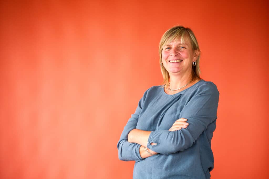 Smiling woman with arms crossed against an orange background, wearing a blue top.