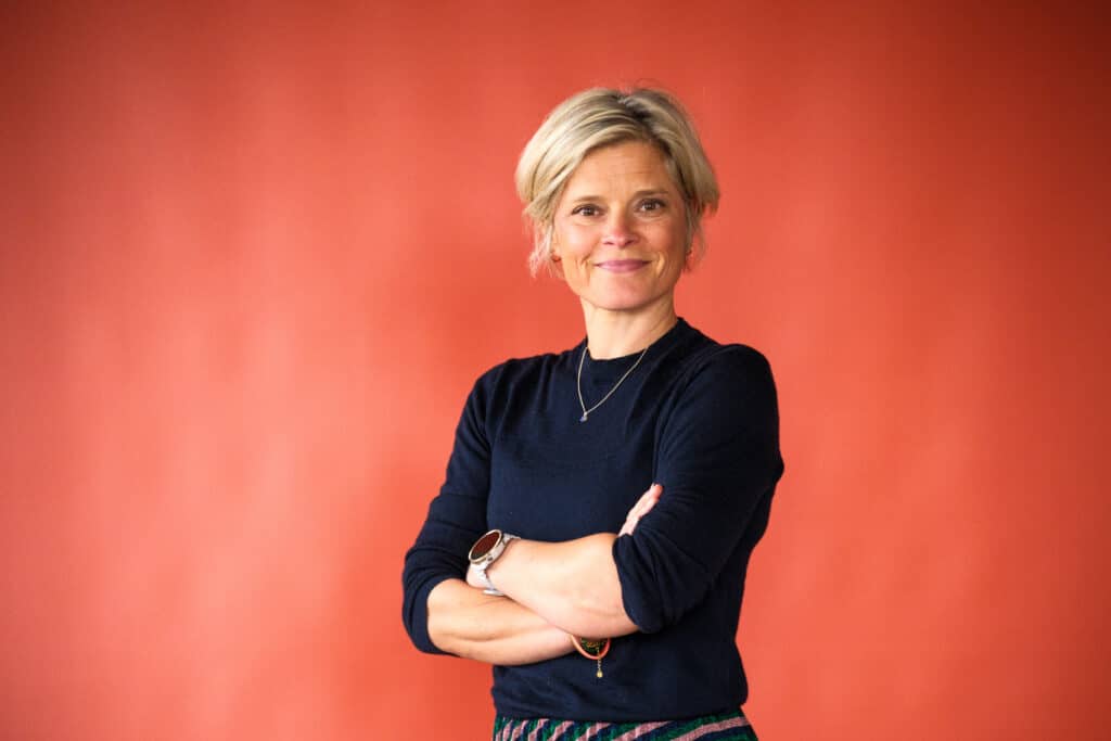 Woman with short blonde hair, wearing a navy sweater, arms crossed and smiling against an orange wall.