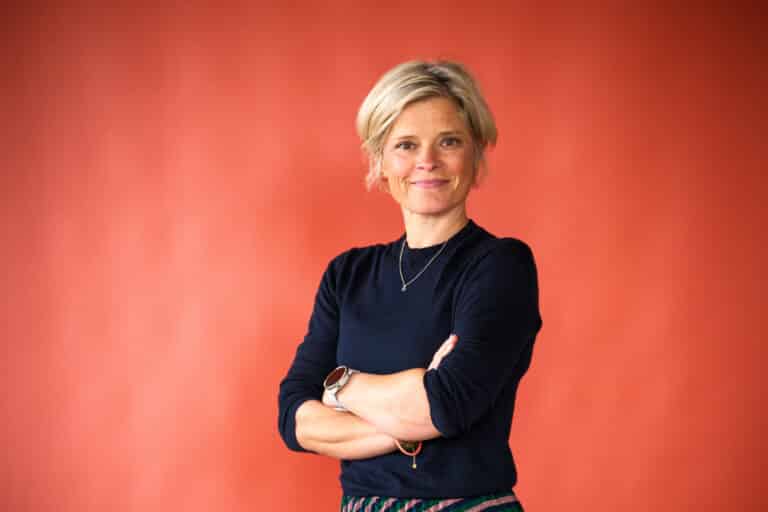 Woman with short blonde hair, wearing a navy sweater, arms crossed and smiling against an orange wall.