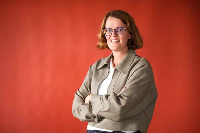 Woman with short brown hair and round blue glasses smiles, arms crossed against an orange background.