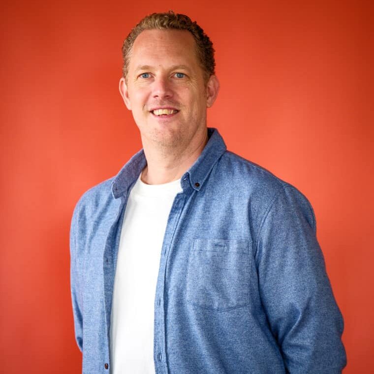 Smiling man in a white t-shirt and blue denim shirt, posing against an orange wall.
