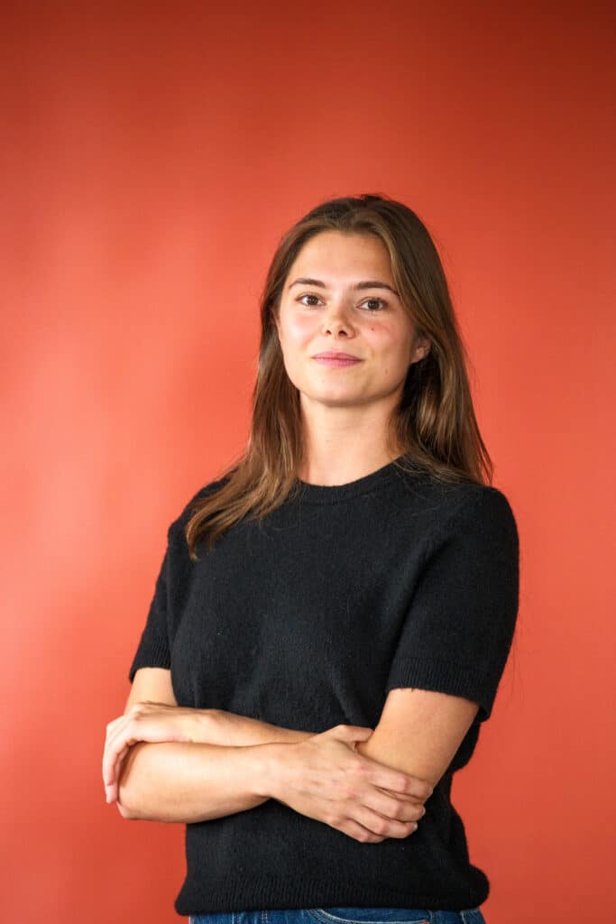 Portrait of a woman with brown hair wearing a black knit sweater, arms crossed, against an orange background.